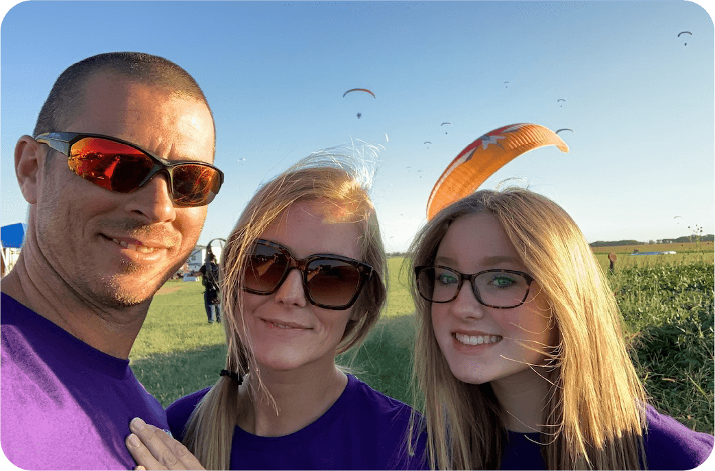 Shawn Elder with his wife Tiffany and daughter Haley at the field, paragliders in the sky behind them.